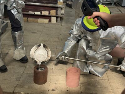 Students wearing protective gear pour molten metal into a mold.