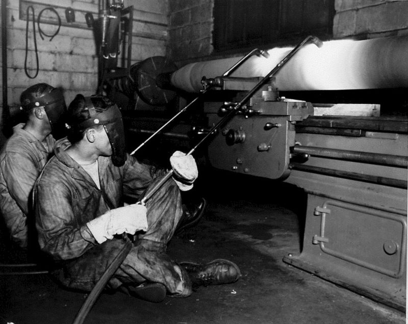 Harold Watson, back, founder of HFW Industries Inc., fuses a thermal spray coating to a large metal shaft. HFW Industries still uses the same spray and fuse process today.