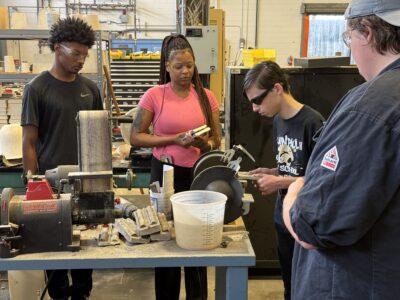 A student polishing a cast piece of metal on a grinder while others wait their turn.