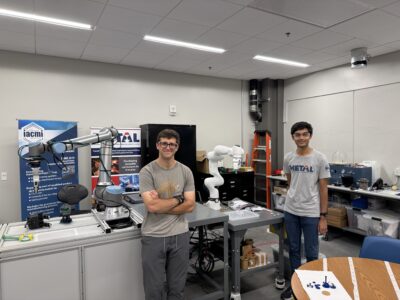 Two students pose with a robotic arm in a laboratory setting. 